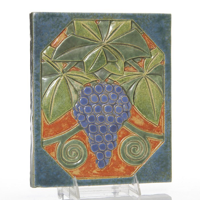 Flint Faience tile, grapes, 8 3/8” x 10”: Flint Faience tile with a grape design, covered in green, blue, purple and orange mat glazes. Unmarked. The lower left hand corner was chipped prior to firing and there is a tiny unobtrusive nick on t