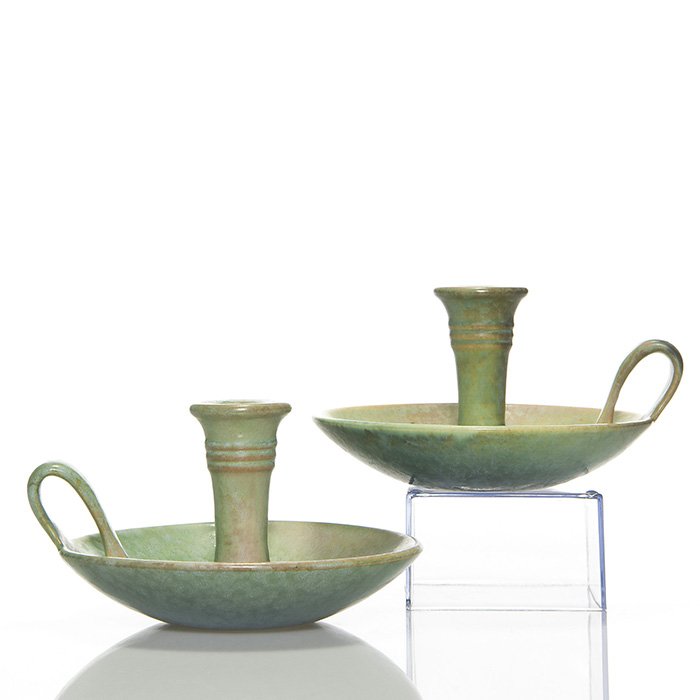 Pr. Roseville Earlam chambersticks, 4”, shape 1080: Pair Earlam chambersticks with loop strap handles. Measures 4 inches by 6 1/2 inches across the tray. One has a black Roseville Pottery sticker and patches of cobalt color in the glaze. Faintly marked