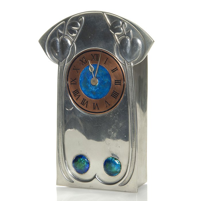 Liberty Tudric clock, enamel, brass dial, 8”: Good Archibald Knox, Liberty Tudric Arts & Crafts mantle clock featuring vine design in relief, a brass and blue enamel dial and twin enamel blue cabochons on the front. Measures 8 inches by 5 3/4 inc