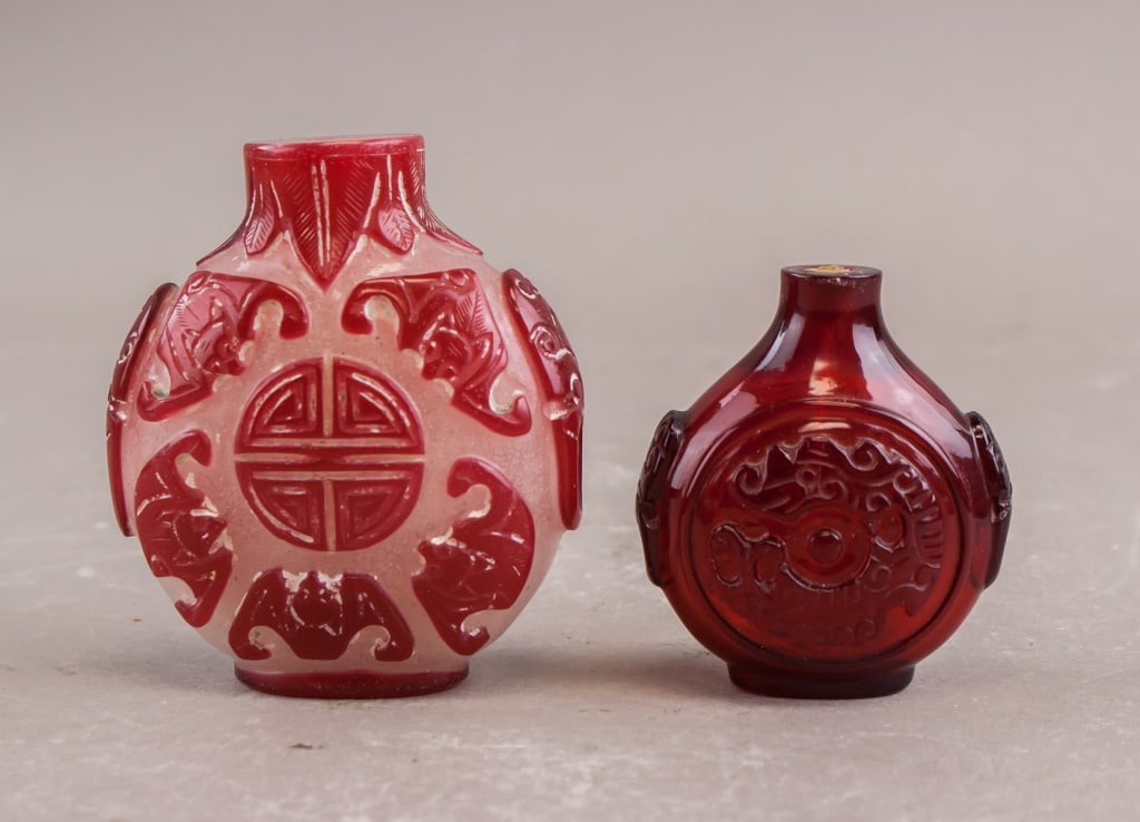 Chinese Red Peking Glass Snuff Bottles 2pc: This lot comprises two Chinese red Peking glass snuff bottles. One bottle is a translucent red with an opaque pink overlay, carved with auspicious motifs and a central medallion featuring a stylized