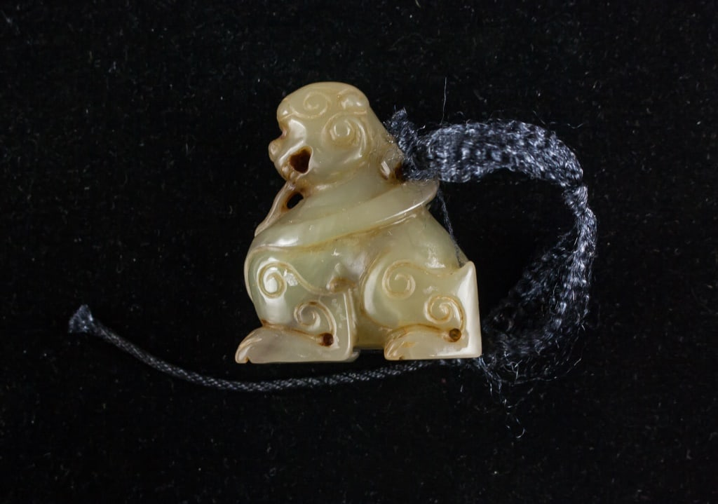 Chinese Han Style Old Jade Carved Lion Pendant: Chinese Han style old jade pendant, carved in the form of a recumbent lion with a curled tail and stylized mane. The jade displays a mottled beige and russet coloration with areas of calcification and