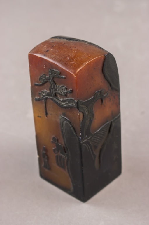 Chinese Brown Shoushan Stone Carved Seal: A Chinese Shoushan stone seal carved in relief with a landscape scene showing figures, a bridge, and a tree, the material displaying a natural transition from brown to darker tones toward the base. H: