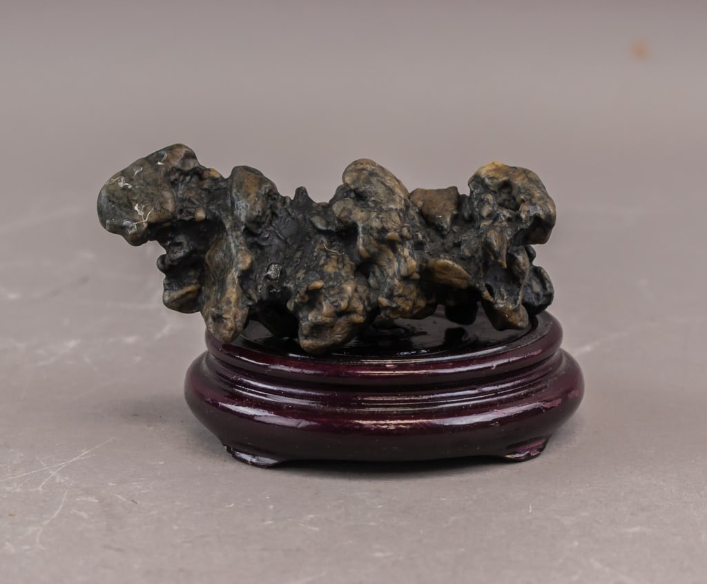 Chinese Inkstone Scholar's Rock w/ Stand: A Chinese scholar’s rock of dark, irregular form, displaying natural surface texture and colour variation, offered together with a fitted wooden stand. The stone shows prominent ridges and cavities 