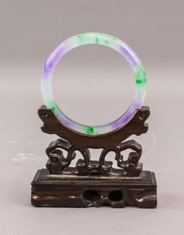 Chinese Green & Lavender Jade Bangle: A Chinese jade bangle of circular form displaying mixed green and lavender tones, well-polished and translucent with natural colour variation throughout. The piece is presented on a carved wooden stan