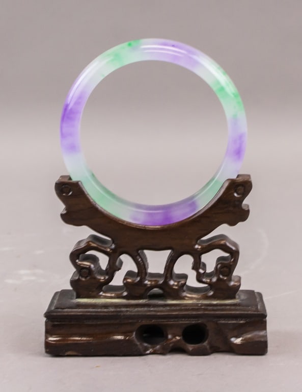 Chinese Green & Lavender Jade Bangle: A Chinese jade bangle of circular form displaying mixed green and lavender tones, well-polished and translucent with natural colour variation throughout. The piece is presented on a carved wooden stan
