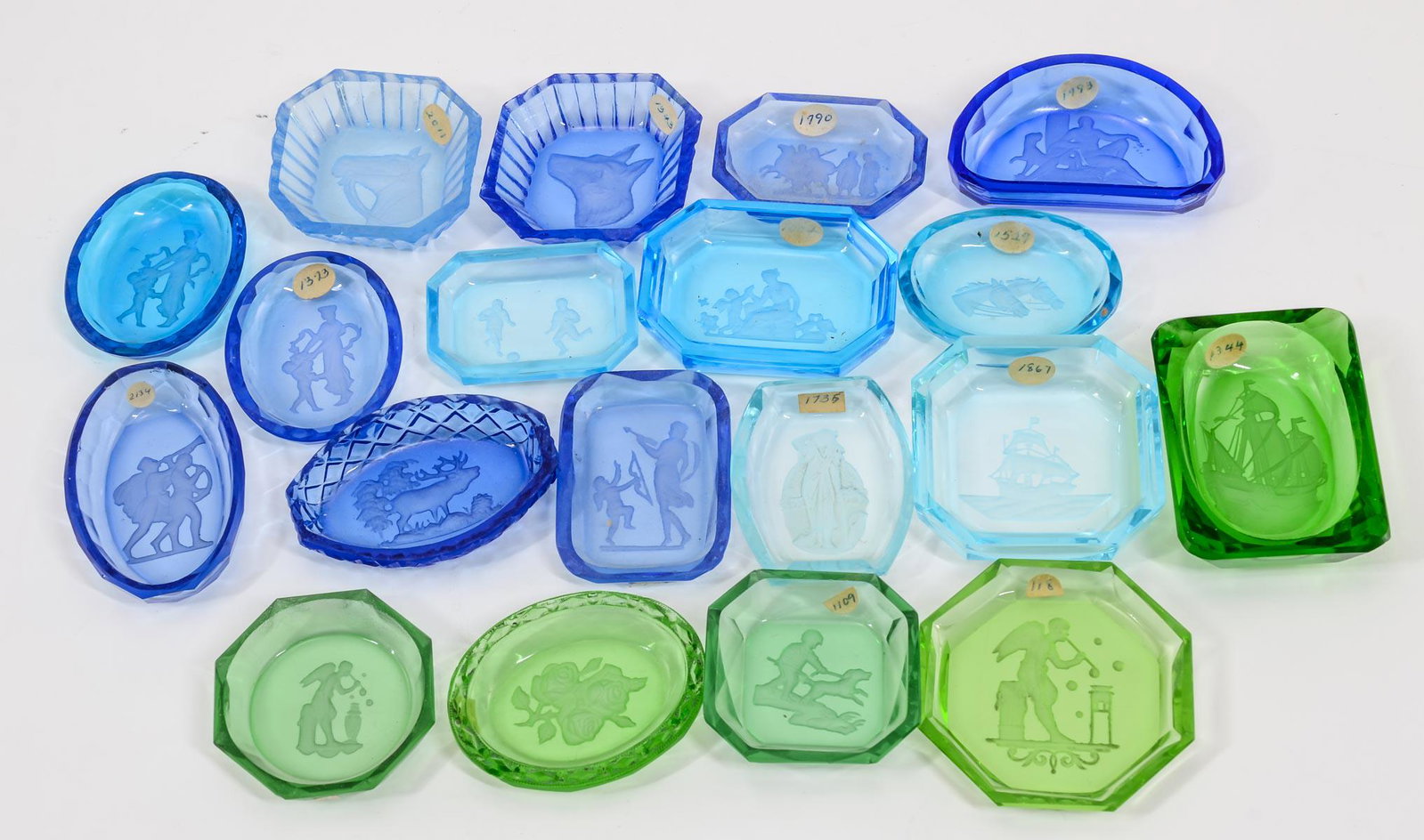 19 Intaglio Glass Open Salts: Nineteen Intaglio cut open salts. 14 in shades of blue, 5 in shades of green. Subjects include: Floral, boy with dog, Angels, ship, Classical scenes, dog head, etc. All in very good condition, except 