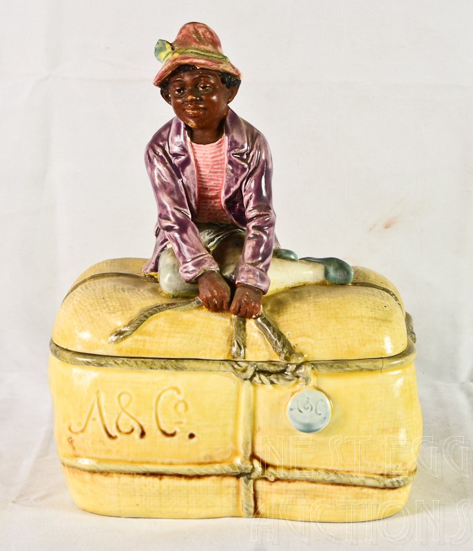 Figural Black Americana Majolica Tobacco Humidor: This rare tobacco or cigar jar depicts a Black boy sitting on top of a tied package or bale. Marked on the side: A & Co. 8.5" x 6" x 4.5" Fine crazing, a very tiny underglaze line o