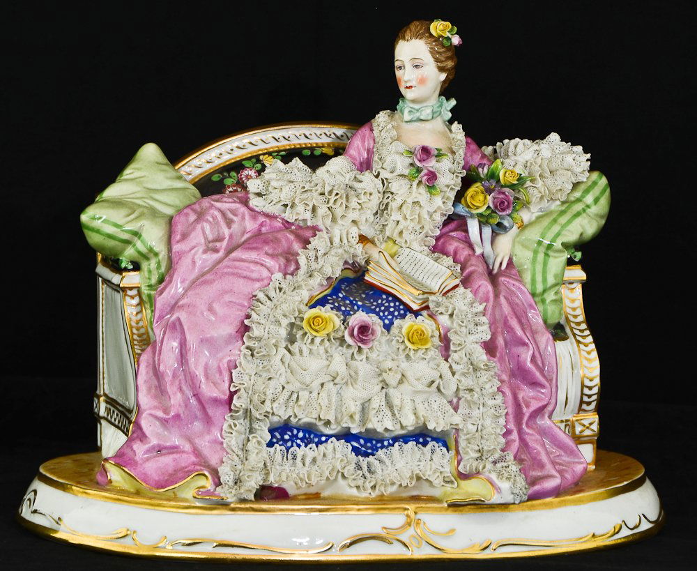 Italian Capodimonte porcelain reclining female figurine: Marked with "Crown N" hallmark on the underside in blue. Crown has five points. Piece features female figure reclining on a loveseat, reading a book. Minor losses to the lace near the base and by her