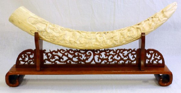 CHINESE IVORY TUSK OF 18 IMMORTALS HAND CARVED: Tusk depicts 18 immortals with a dragon and trees. Has fitted wooden base. 20th Century. No apparent signature. Measures 25" L (64cm).