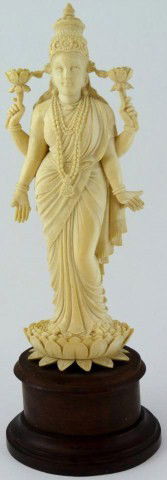 ANTIQUE INDIAN IVORY TRIDEVI GODDESS FIGURE: Hand carved Indian ivory Trivedi Goddess figure. She is wearing layered robe with etched trim decoration. In two hands she is holding lotus blossoms and she is standing on a large lotus blossom. Mount