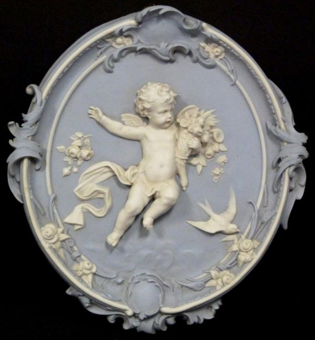 ANTIQUE GERMAN BISQUE CHERUB WALL PLAQUE: Antique German light blue and white bisque porcelain wall plaque. Has figural cherub, bird and floral design with scrolled border embellishments. 19th century. Measures 16 1/2" height x 15" width (41.