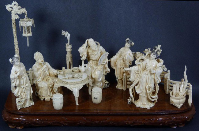 CHINESE CARVED IVORY PARLOR SCENE GROUP: Chinese hand carved ivory group figure. Depicts a parlor scene with 3 maidens. An old man is sitting at a table and there is a man helping him. Also in the scene is garden seats, lamp post and lantern
