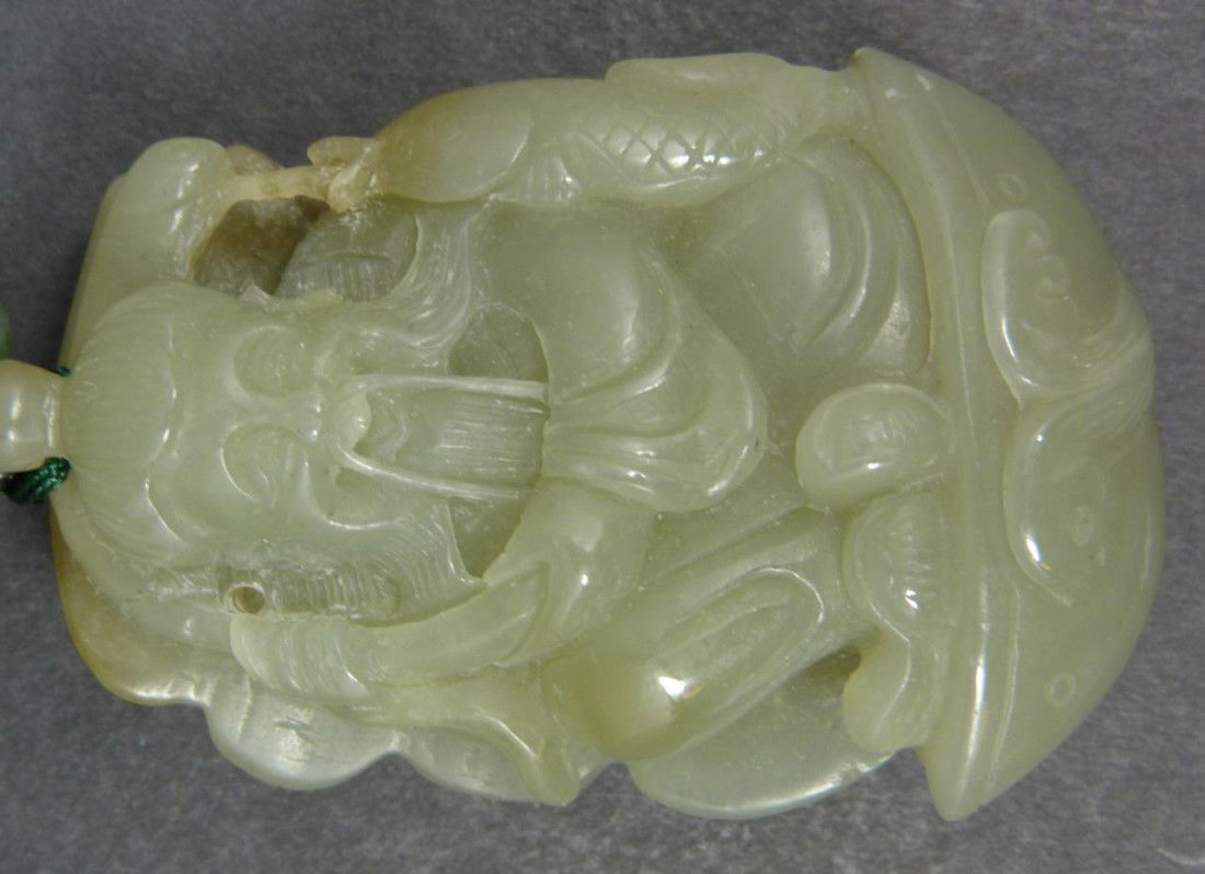 HAND CARVED CELADON JADE FISHERMAN IN SAMPAN: Hand carved celadon jade toggle figure depicting a fisherman in sampan boat. Finely carved with beautiful translucence throughout. Some pebbling to sail with hints of brown jade. Has woven wrist strap