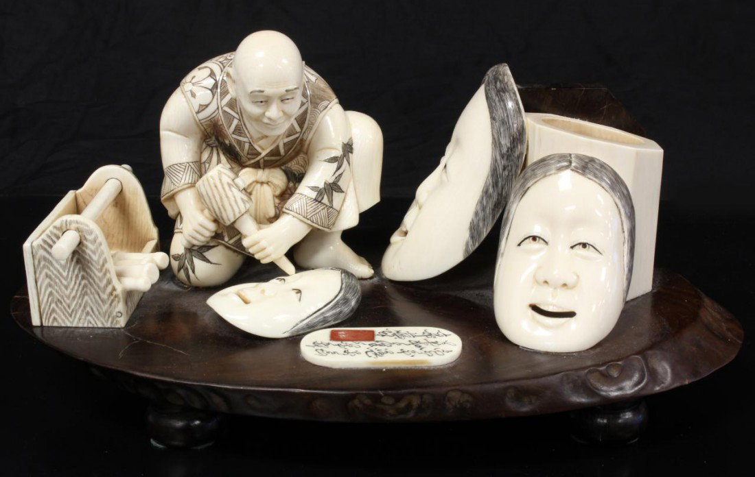 JAPANESE CARVED IVORY FIGURE OF MASK MAKER SIGNED: Japanese ivory and wood figure depicting a mask maker diligently at work. He is kneeling over a mask as he chisels away on it. Next to him is his tool box and finished masks. Signed to plaque on base.