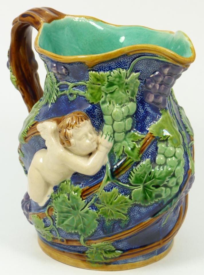 MINTON MAJOLICA PITCHER GRAPE VINE AND CHERIBIM: Minton, earthenware pitcher, deep navy, green, purple and brown slip, depicting a grape vine encircling the piece, highlighted with conforming winged cherubim. The brown branch design handle has a pur