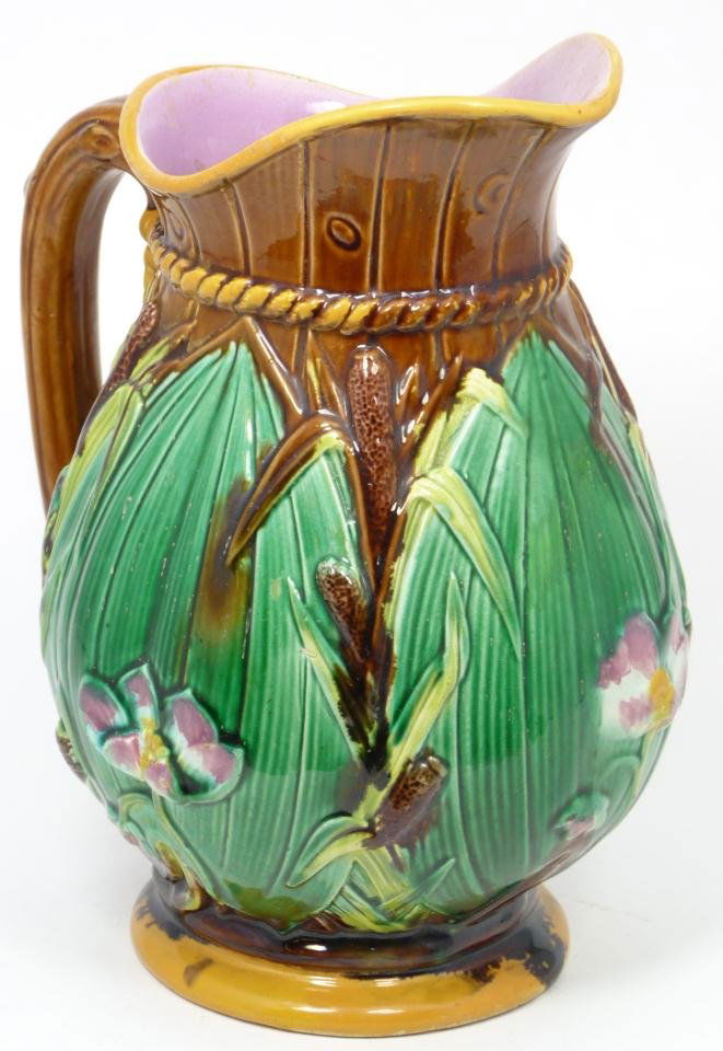 MAJOLICA POTTERY PITCHER CATTAIL AND FLORAL: Majolica, earthenware pitcher, emerald green and brown slip, cattail, foliage and floral pattern, with lavender interior and brown branch handle. Marked 1800 M on the bottom of the footed base. Measur