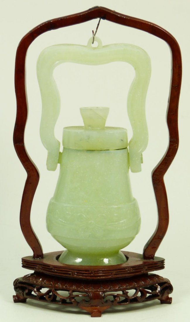 CHINESE CARVED JADE HANDING URN AND STAND: Chinese hand carved jade hanging urn having raised floral band to body and hinged handle. Has fitted wood display stand. Pale green translucent mottled jade specimen. 20th century. Measures 6 1/4" hei
