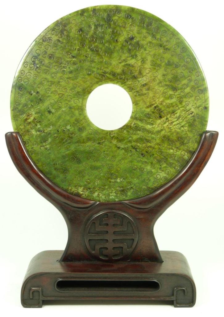 CHINESE CARVED SPINACH JADE PI DISC WITH STAND: Chinese carved spinach jade bi disc having etched swirl designs to one side. Verso having etched cross T design. Fine thin carving. 19th/20th century. Disc measures 10" diameter + 5" base height (25.4