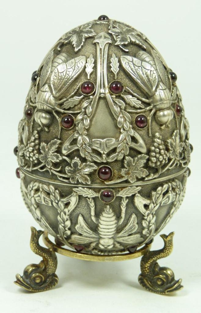 RUSSIAN SILVER JEWELED FLIES EGG: Russian silver egg. Has intricate raised design depicting flies, wreaths and grape clusters. Mounted with Cabochon garnets throughout. Gold wash interior. Has Pavel Ovchinnikov workmaster marks to int