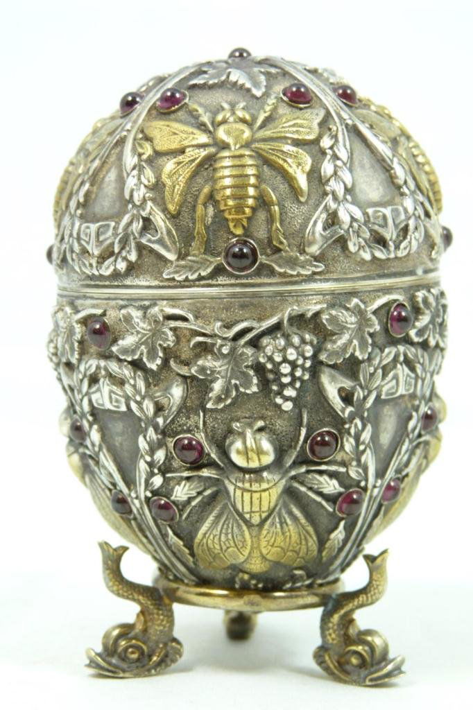 RUSSIAN SILVER JEWELED FLIES EGG: Russian silver egg. Has intricate raised design depicting flies, wreaths and grape clusters. Mounted with Cabochon garnets throughout. Gold wash interior and insects. Has Pavel Ovchinnikov workmaster