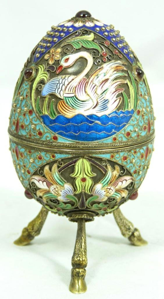 RUSSIAN SILVER FIGURAL JEWELED FOOTED EGG BOX: A Russian silver enameled egg box depicting swans and flowers with filigree and beaded design throughout. Holds cabochon ruby to top with three rubies set to base. Has tripod goat hoof feet. Gold wash