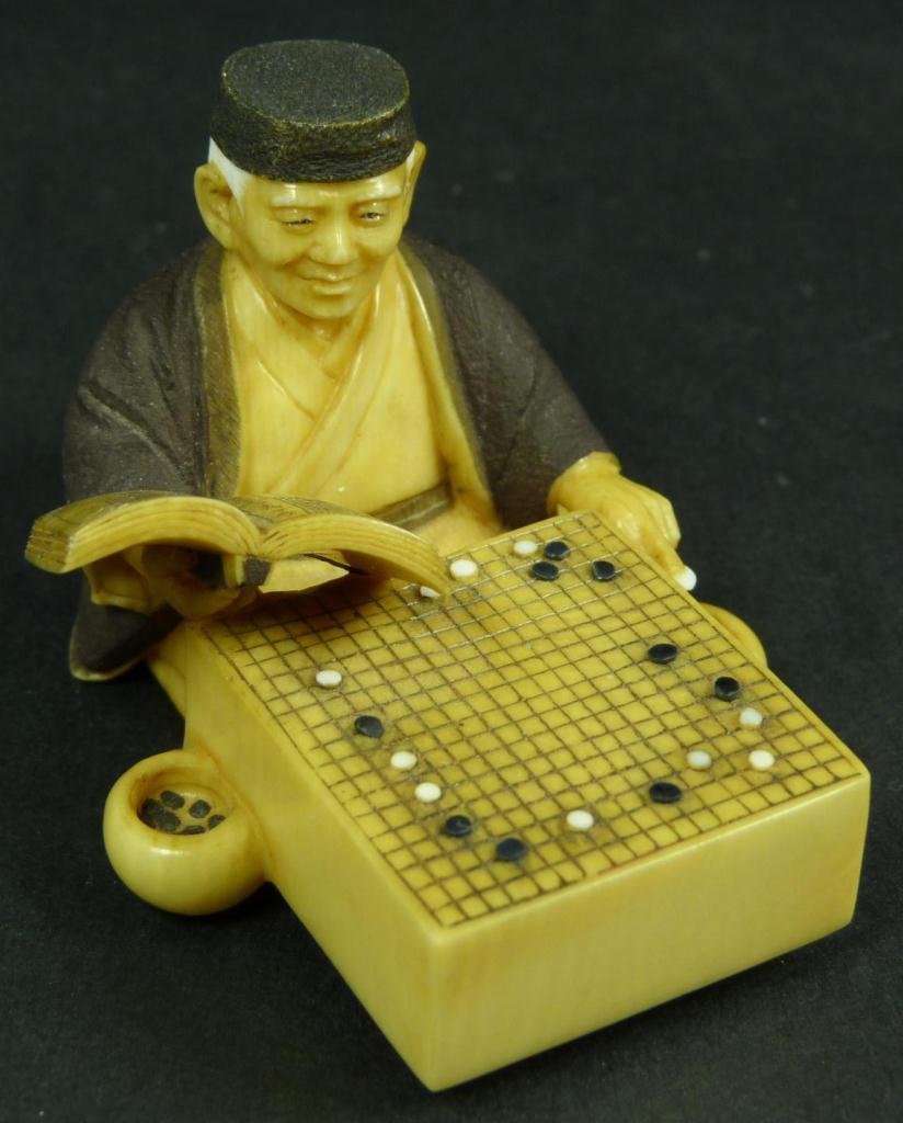 FINE JAPANESE CARVED IVORY NETSUKE SIGNED KOYU: An exquisitely carved Japanese ivory netsuke figure depicting a wiseman studying games of go. Signed Kuyo to bottom on red plaque. Includes wooden presentation box. Circa 1965-1970. Finely carved and 