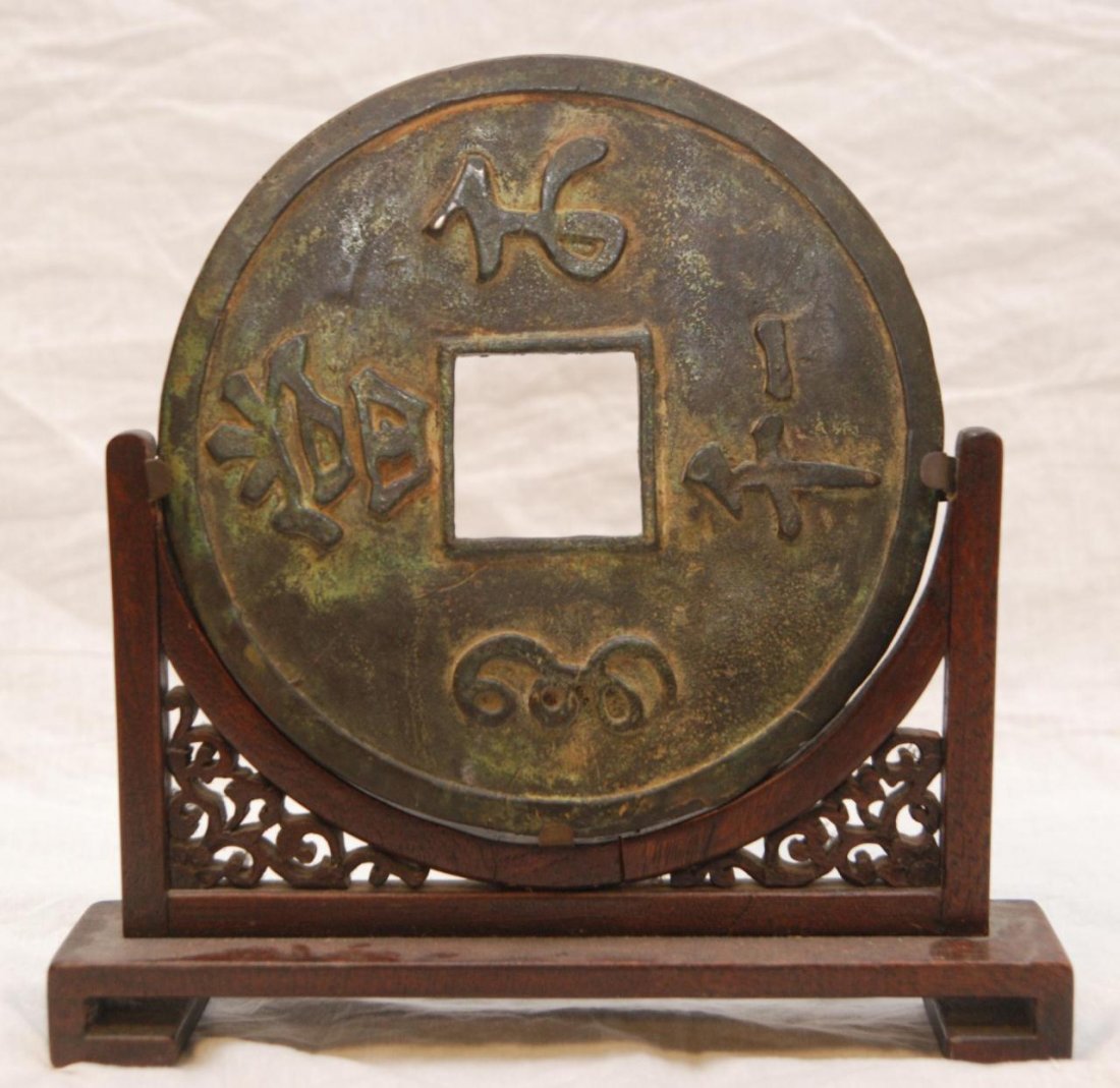 CHINESE LARGE CAST BRONZE CAST TREASURY COIN: Chinese large cast bronze treasury coin. Depicts calligraphy to front and back. Includes fitted wooden display stand. Measures 7 5/8" diameter x 3/8" depth + 2" base height (19.3cm x .9cm + 5.1cm). To