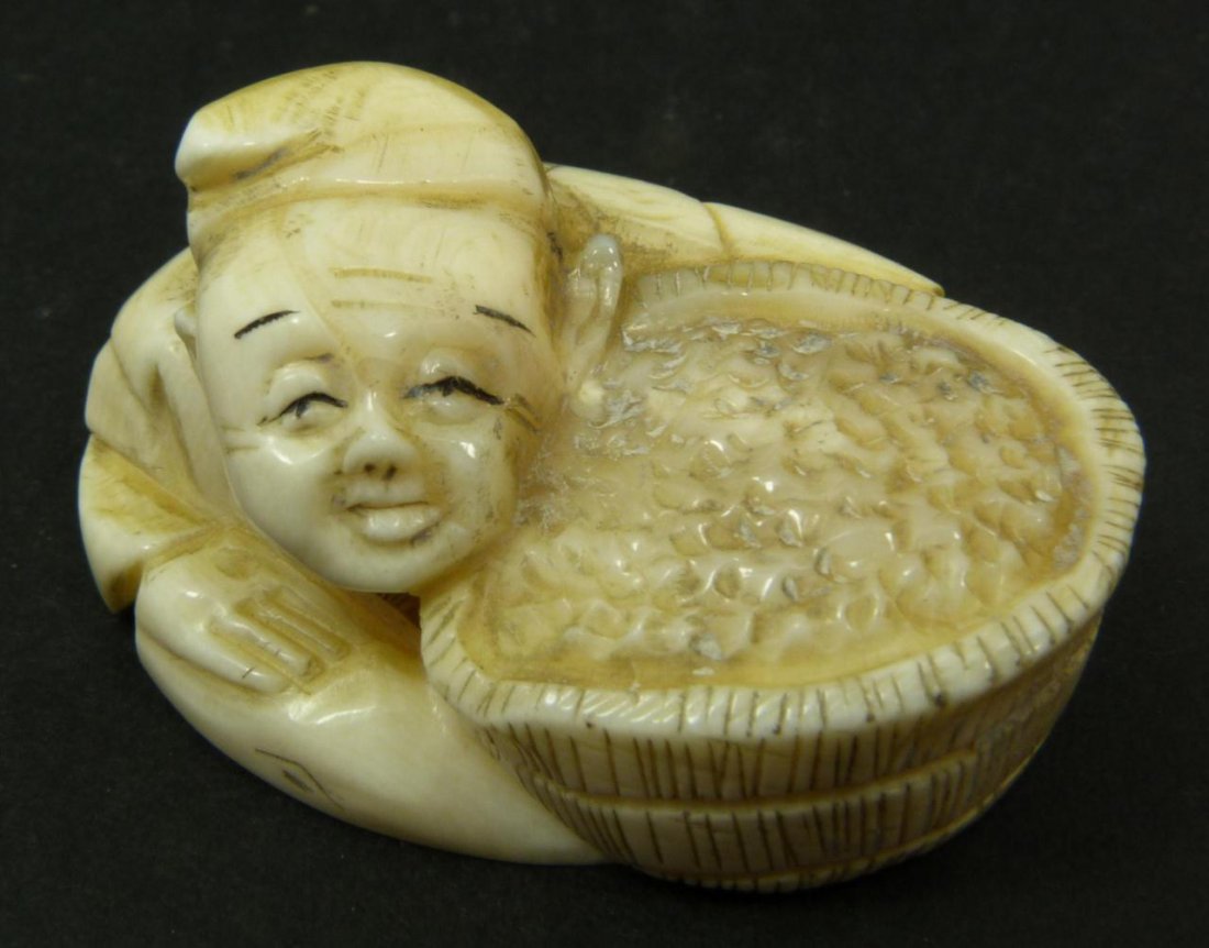 JAPANESE CARVED IVORY NETSUKE OF BOY WITH GRAIN: Japanese hand carved ivory netsuke depicting a seated boy with basket of grain. Etched calligraphy signature to bottom. Measures 1in Height x 2in Width. Weighs 34g.