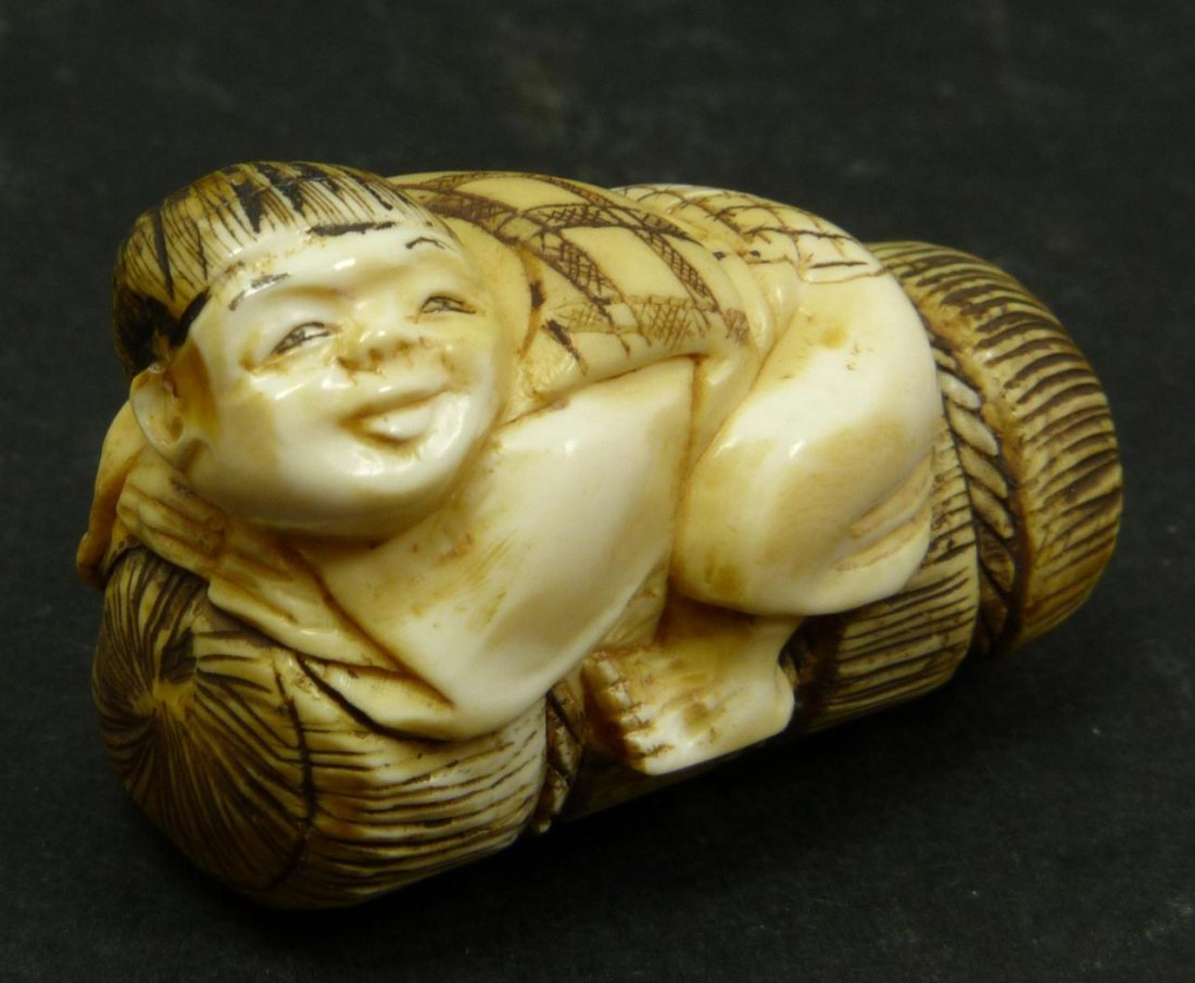 JAPANESE CARVED IVORY NETSUKE OF CHILD: Japanese carved ivory netsuke figure depicting a child on bindle. Etched calligraphy signature to bottom. Measures 1 7/8in Length, Weighs 26g.
