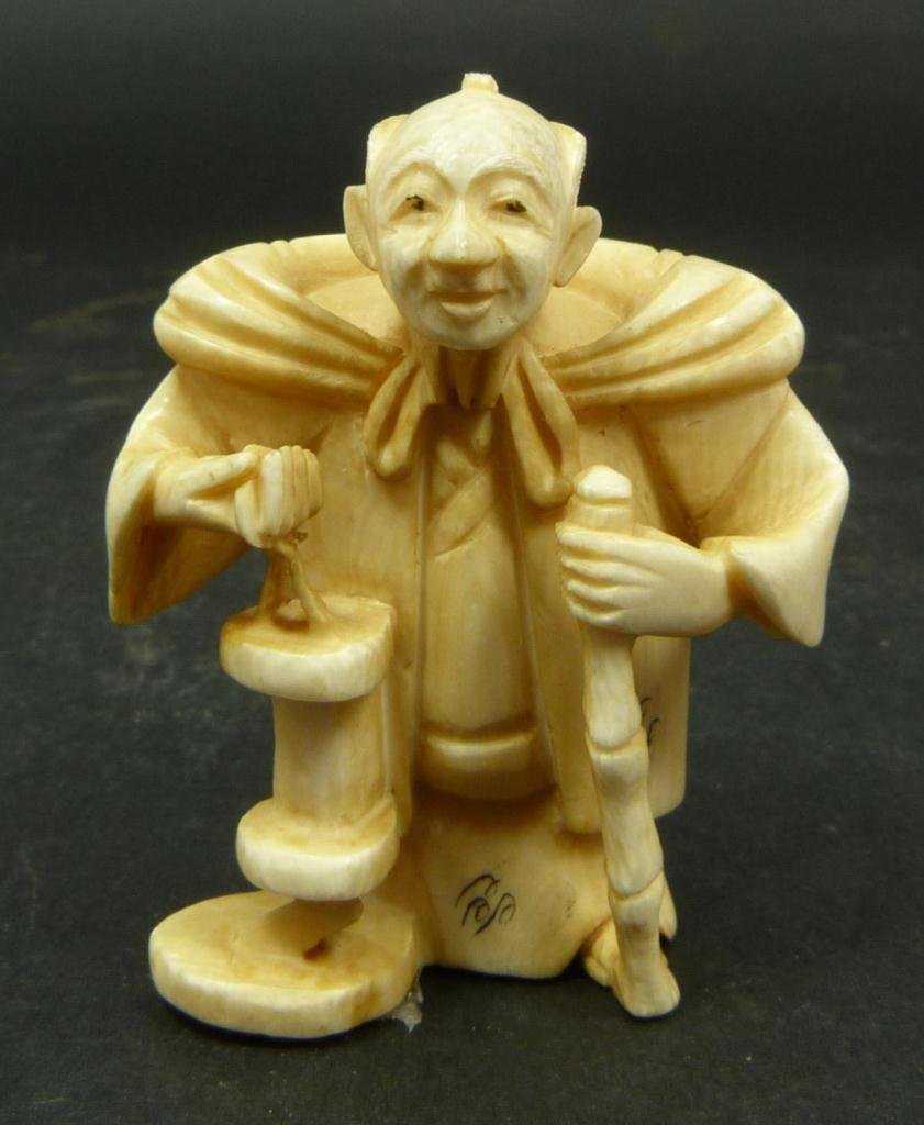 JAPANESE CARVED IVORY NETSUKE OF MAN WITH LANTERN: Japanese carved ivory netsuke figure depicting a standing man with bamboo cane and lantern. Etched calligraphy signature to bottom. Measures 2in Height. Weighs 23g.