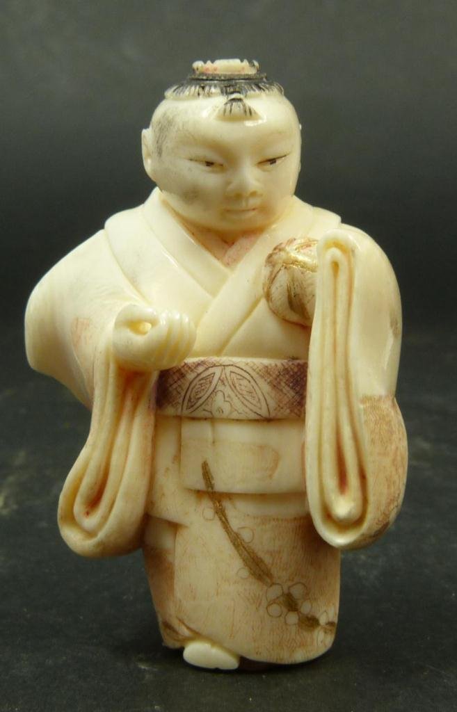 JAPANESE CARVED POLYCHROMED IVORY NETSUKE OF CHILD: Japanese carved and polychromed ivory netsuke figure depicting a boy in layered robe holding a ball. Etched gilded calligraphy to bottom. Measures 2in (5.1cm) Height. Weighs 28g.