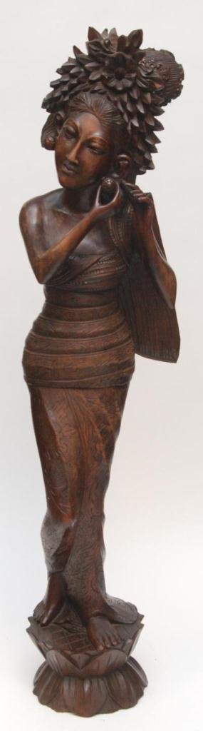 BALINESE CARVED WOODEN SCULPTURE OF MAIDEN: A hand carved Balinese carved wood sculpture depicting a standing maiden in wrap dress putting on peg earrings. She is standing on stylized lotus blossom base. Measures 29" height (73.6cm).