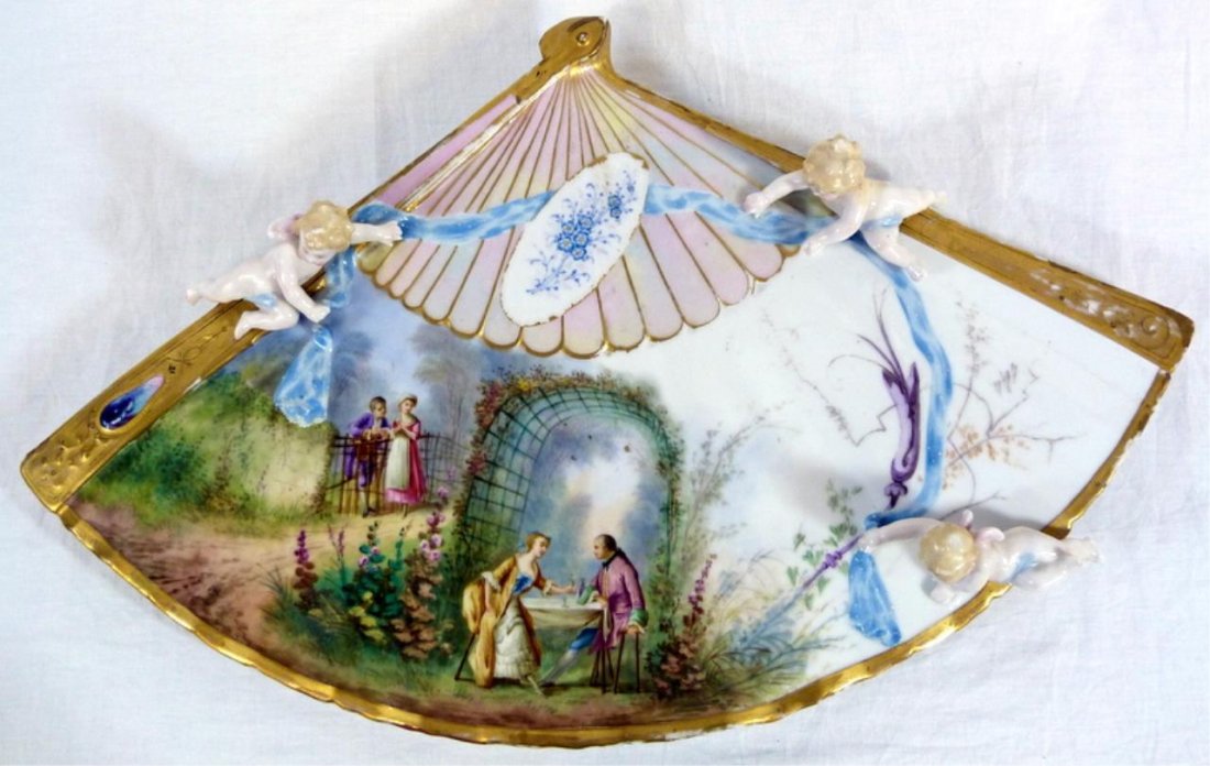 FRENCH PORCELAIN FIGURAL FAN & CHERUBS PLAQUE: Antique French porcelain hand painted fan form wall plaque having three figural cherubs holding blue ribbon. A scene of a courting couple as children can be seen to left, the center depicts the couple