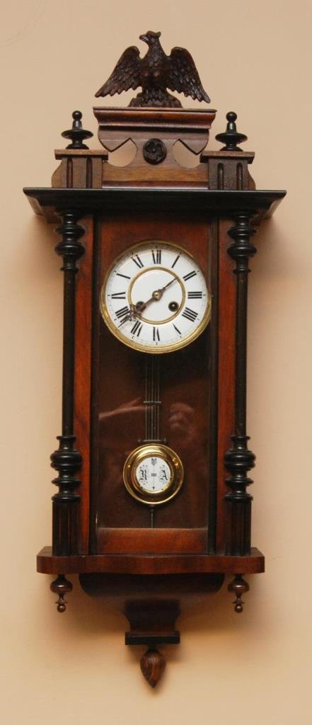 KINZLE SCHWENNINGEN WOODEN CASE WALL CLOCK: Antique Kinzle Schwenningen German wooden case wall clock having an eagle finial to top and column design to sides. Glass screen to front. White enamel face with black Roman Numerals and gilded brass 
