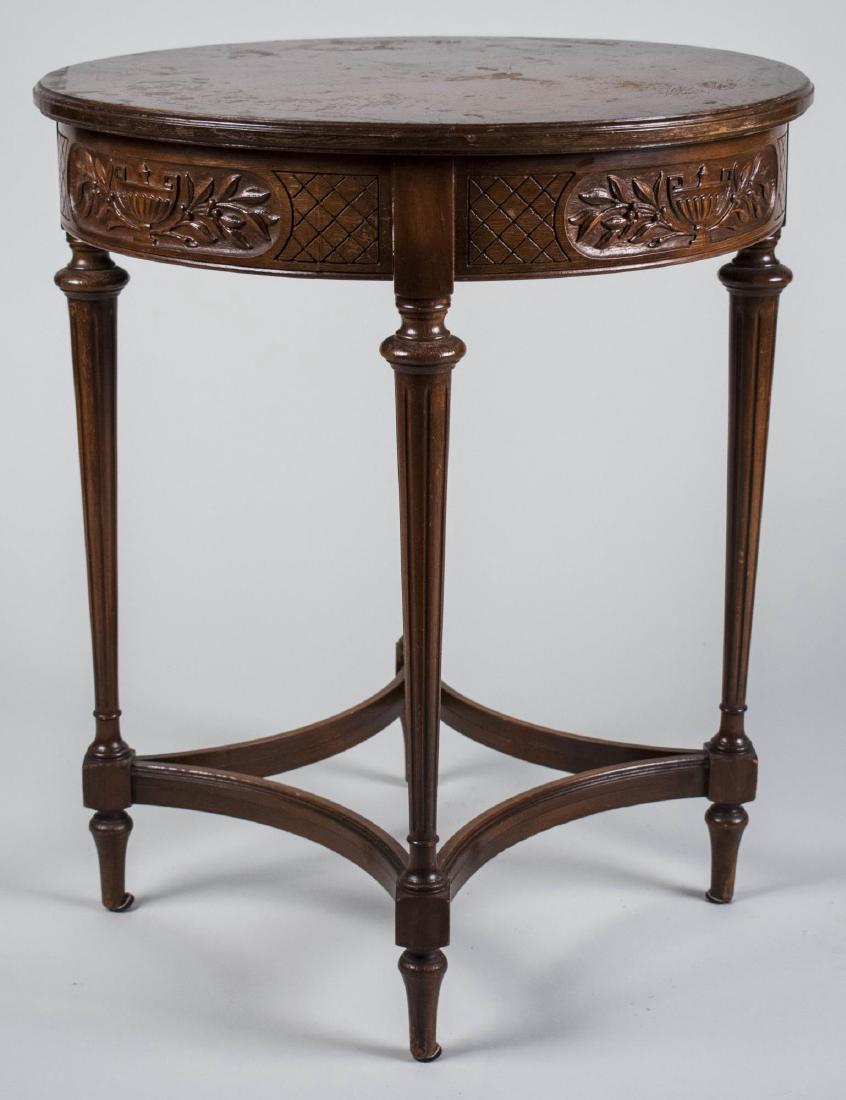 Neoclassical Style Occasional Table: Circular top with crossbanded edge, over a carved frieze, tapered fluted legs joined by a modified box stretcher. Height 28 inches, diameter 24 inches. Notice to bidders: Condition reports and additio