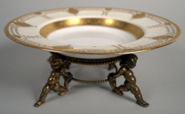 Minton Porcelain Centerpiece: Shallow porcelain bowl with raised neoclassical style gilt decoration, on a gilt metal base with four putti legs. Height 3 1/2 inches, diameter 9 inches.