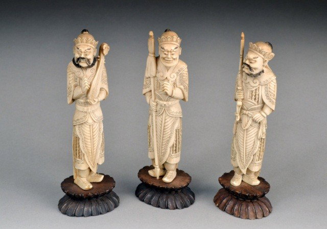 Three Chinese Ivory Guardian Figures: One holding a spear, one wielding a sword, and one carrying an instrument. Each on a wooden stand, tallest height 5 3/8 inches.