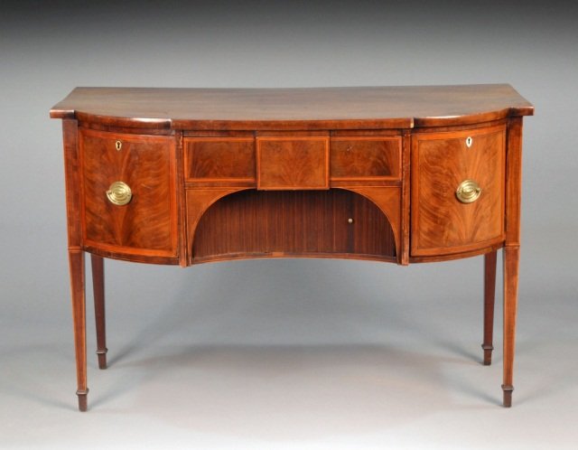 George III Style Mahogany Sideboard: Early 19th century, shaped rectangular top above a central drawer and tambour front storage bin, flanked by a conforming single door cupboard and a deep drawer, square tapered legs ending in spade fee