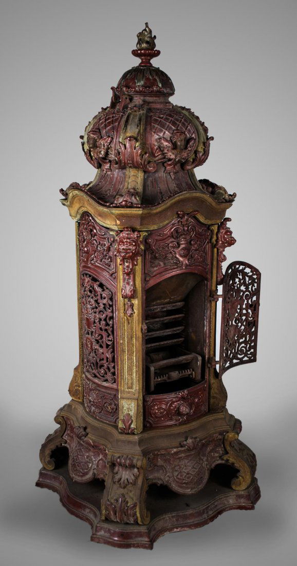 Cast Iron Stove: Deep red painted stove with pierced scroll decoration, domed top with flamiform finial, winged mask accents. Height 54 inches, width 24 inches, depth 24 inches