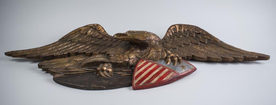Carved Wood Eagle: Modeled with spread wings, holding arrows and a shield with his talons. 14 x 44 inches. Notice to bidders: Condition reports and additional photographs are provided by request as a courtesy to our cli