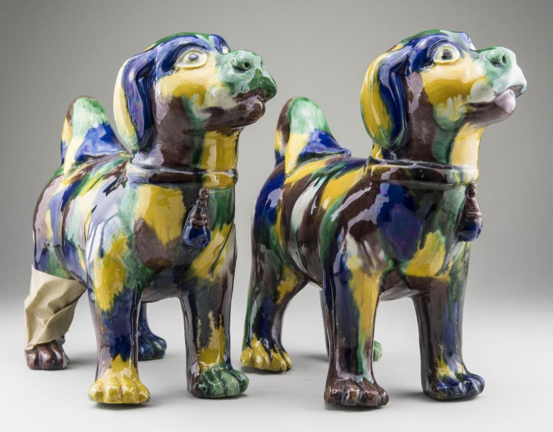Pair of Ceramic Dog Figures: Each modeled standing with a multi colored glaze. Height 7 1/2 inches. Notice to bidders: Condition reports and additional photographs are provided by request as a courtesy to our clients, as such any
