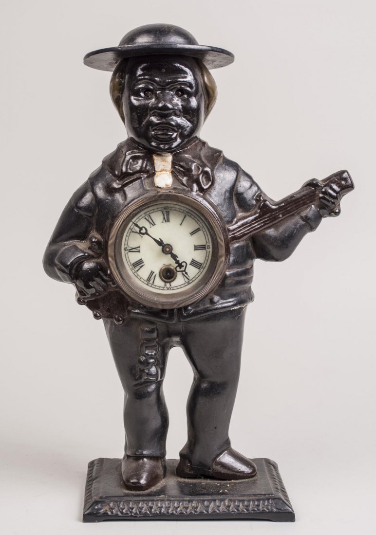 Cast Iron Black Musician Figural Clock: Modeled as an African American man standing playing a banjo, enamel face with Roman Numeral chapters and Arabic minutes. Height 15 inches. Notice to bidders: Condition reports and additional photograp