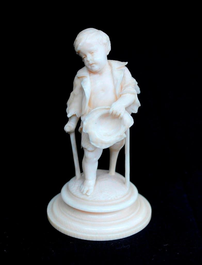 Carved Ivory Figure of a Small Angel: This is a wonderful old carved ivory figure of a small crippled angel boy. The boy is standing barefoot on a crutch while holding his hat in one hand and a cane in the other, his wings come through hi