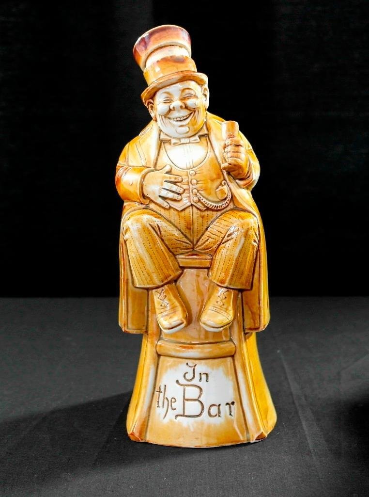 Vintage Schafer and Vater Flask - Brown Figural Nipper: This vintage Schafer and Vater piece is a brown figural nipper and is titled "In the Bar." It depicts a jolly, rotund man sitting on a stool and enjoying a drink with a big grin on his face. The piece
