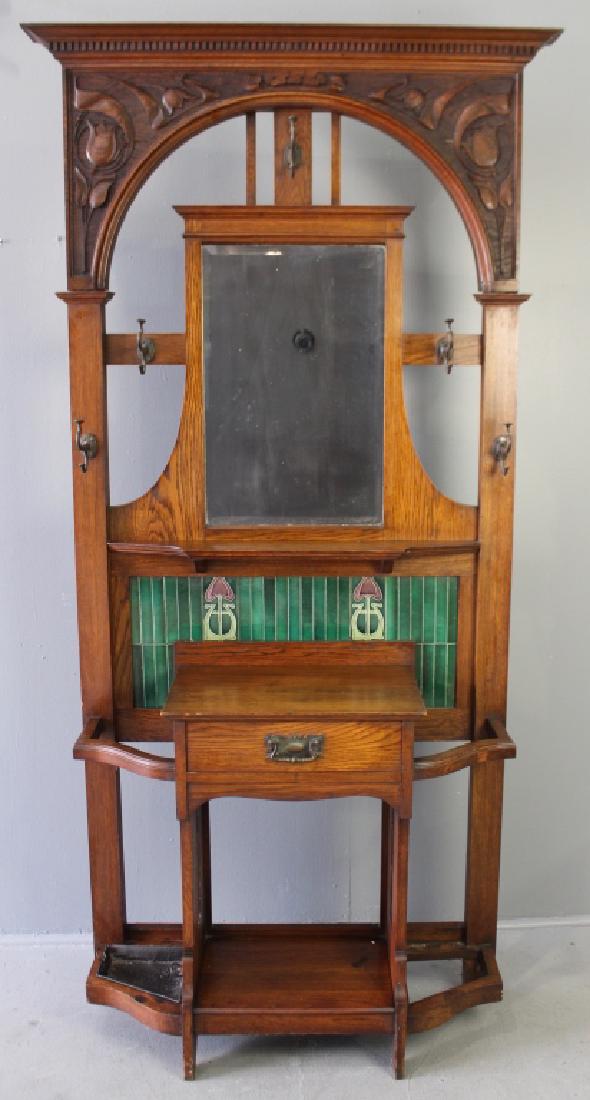 English Arts & Crafts Oak Hall Tree: with tile inset back, beveled mirror, trapezoidal shaped front with single drawer, cut-outs top and bottom, elaborate carved crest with dental molding and overhang top, all original cast iron hangers 
