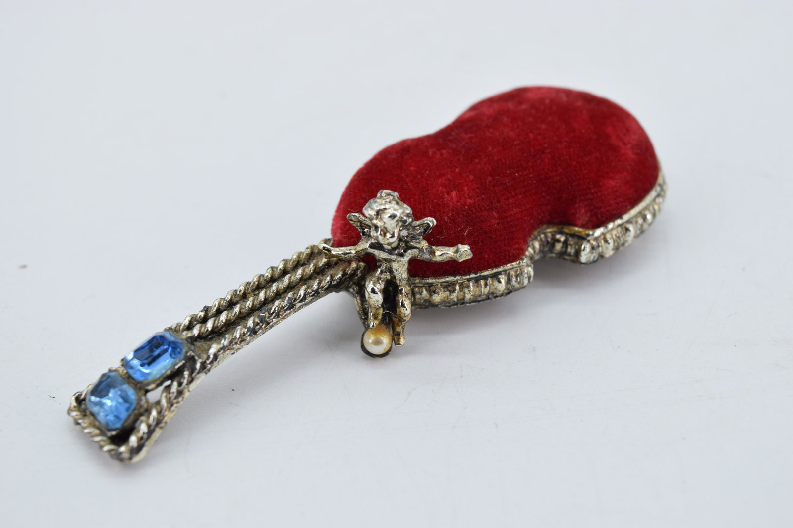 Vintage SF Vanity Pin Cushion with Cherub & Blue Stones: guitar shape with seated cherub with pearl bead and 2 emerald cut faceted blue stones, approx. 4 1/8 inches, red velvet in great condition