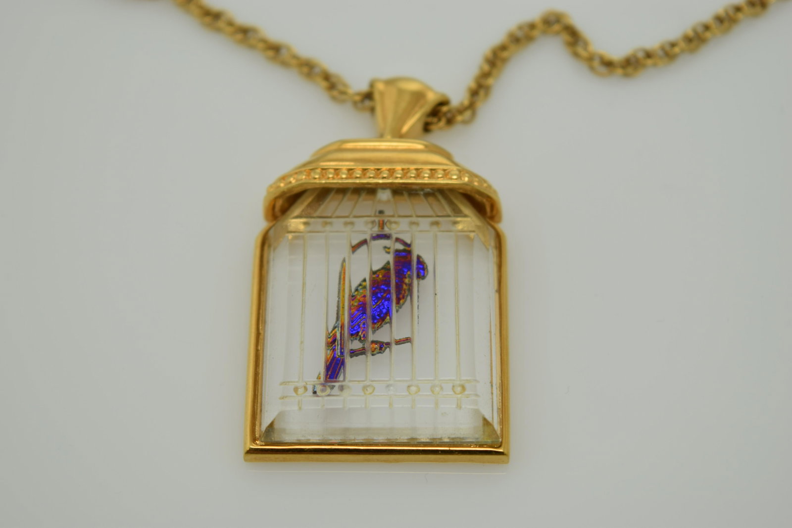 A gold tone chain necklace with a bird cage pendant.: An approx. 30" gold tone chain with a Lucite bird cage pendant with an intaglio iridescent bird. It has a hang tag with a castle on it & is in good condition.
