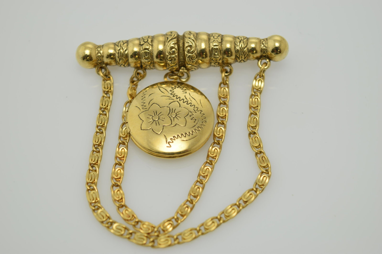 A vintage gold tone locket pin/brooch.: A vintage gold tone pin with 2 chains looped & hanging from the bar section & a gold tone locket hanging in the center with 2 flowers stamped into it. It is in good condition.