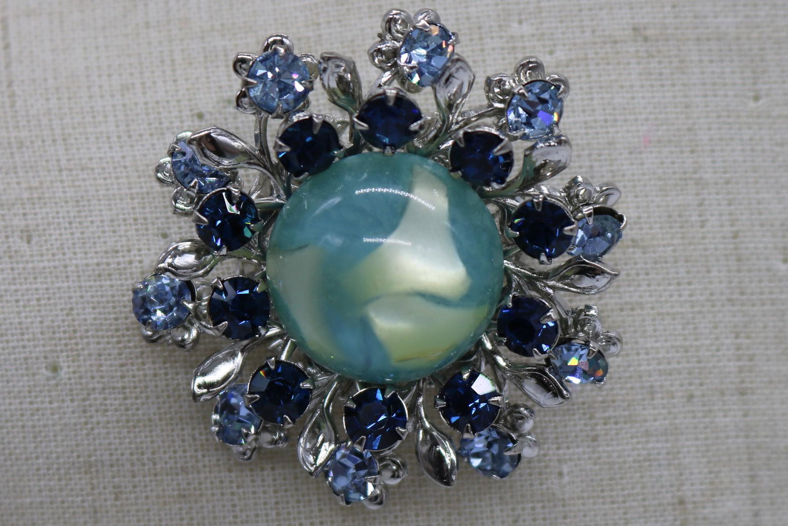 A vintage, round blue rhinestone pin/brooch.: An approx. 1" x 1" round, silver tone pin w a large confetti Lucite center surrounded by 2 shades of blue round rhinestones. It is in good condition.
