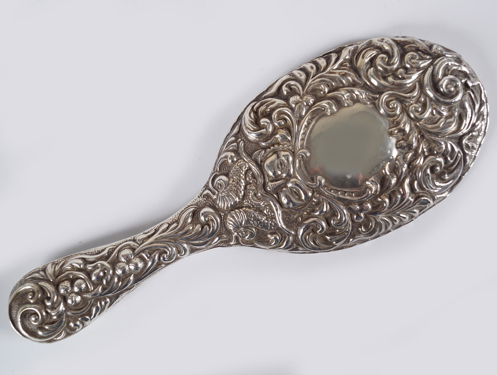 19TH-CENTURY SILVER BACKED VANITY MIRROR: with an oval plate and an embossed mask and a seahorse decorated back and handle.